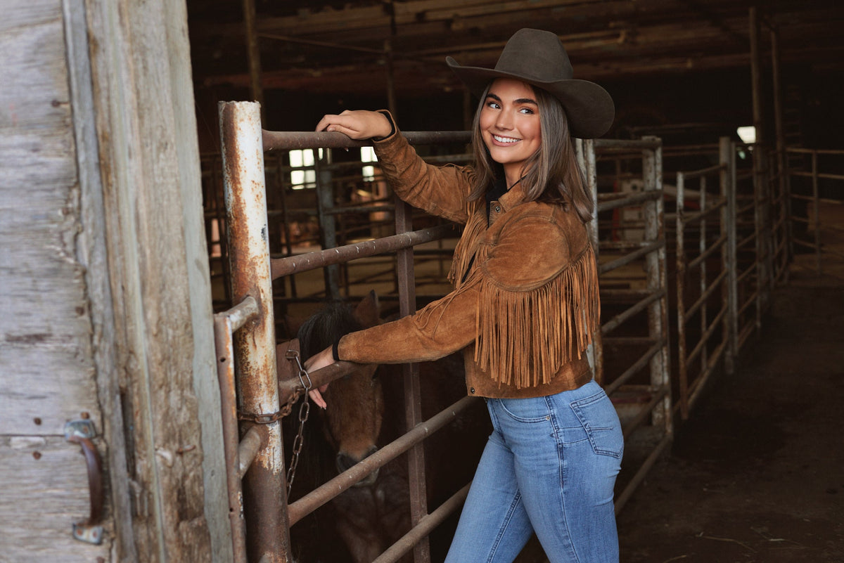Woman in a fringed suede jacket, bootcut jeans and a black felt cowboy hat at a cattle pen