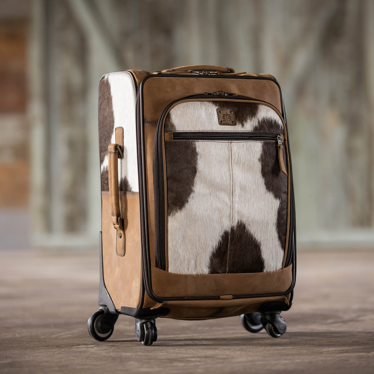 STS Ranchwear cowhide rolling suitcase with sturdy wheels and leather handle, featuring a rugged, natural cowhide exterior.