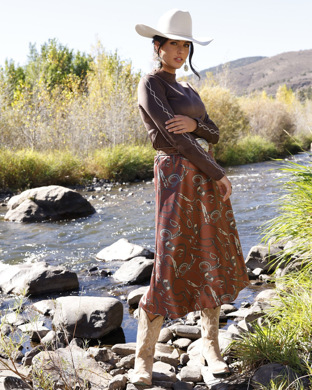 Woman in a brown Cruel Denim embroidered top and western print skirt standing by a mountain river