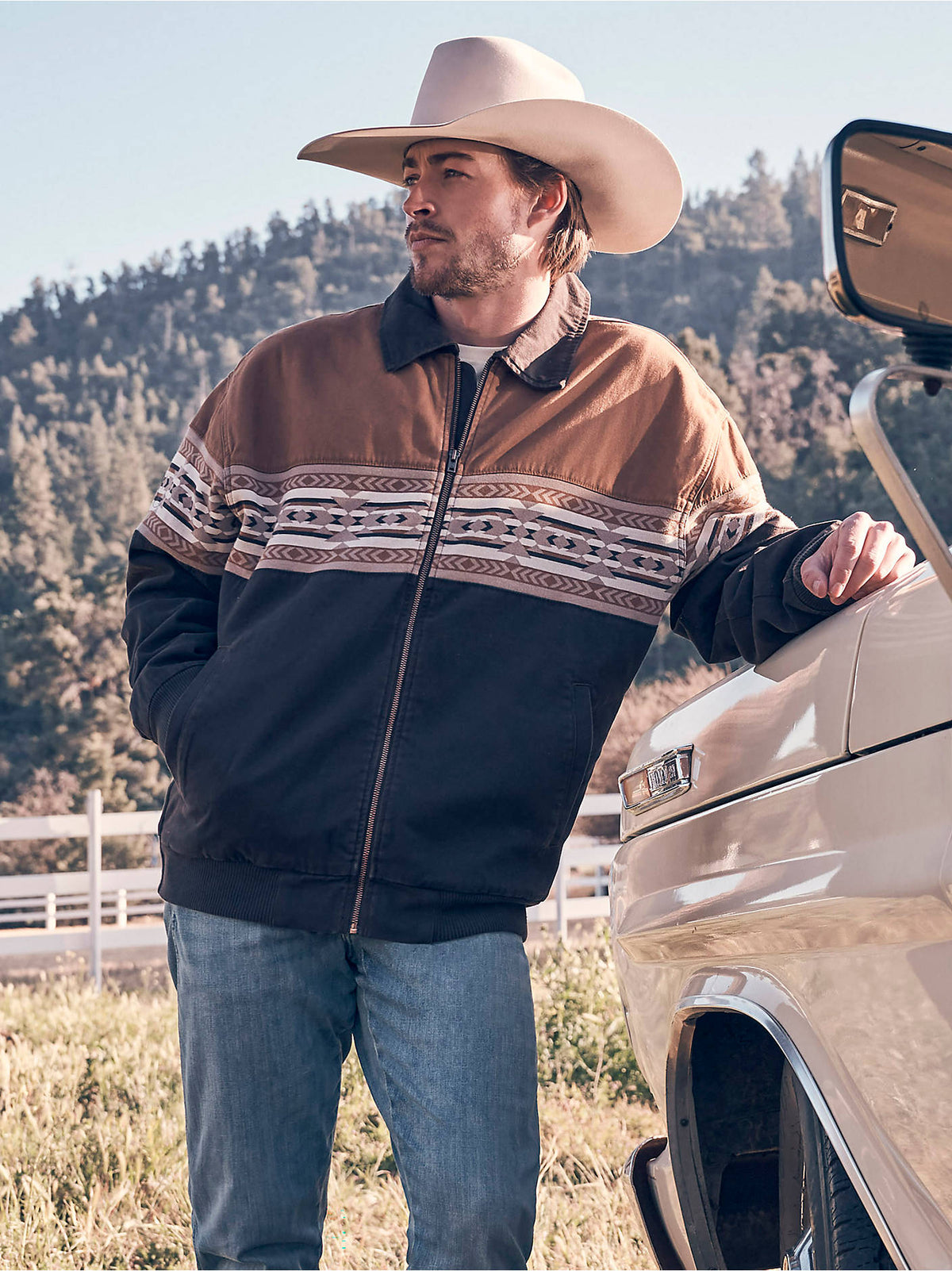 Man wearing the Wrangler® Checotah® Bomber Jacket in Chocolate Torte, a brown and black colorblock bomber jacket with geometric Checotah stripe, leaning against a truck outdoors.