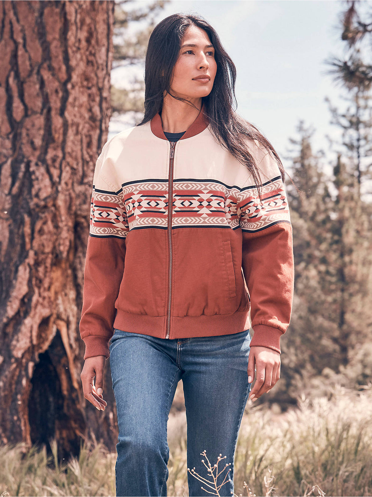 Woman wearing the Wrangler® Checotah® Bomber Jacket in Cream Slice, a cream and rust colorblock bomber jacket with geometric Checotah stripe, outdoors in a forest.